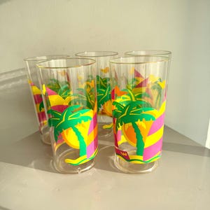 May include: Five clear highball glasses with a colourful tropical design. Each glass features a palm tree with yellow, pink, and green accents. The glasses are arranged on a light-coloured surface.