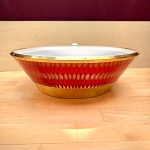 May include: A shallow, circular bowl featuring a vibrant red exterior with a repeating textured honeycomb pattern. The bowl is finished with a polished gold-tone metal rim at the top and a matching gold-tone base, with a smooth white interior.