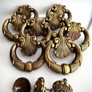 May include: A selection of antique brass drawer pulls and knobs. The drawer pulls feature a shell design with a circular handle. The knobs have a floral pattern. The hardware has a vintage, aged appearance.