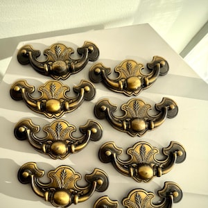 May include: Eight antique brass drawer pulls with a decorative leaf design. The pulls have a curved handle shape and are arranged on a white surface. The brass finish has a vintage look, with dark accents.