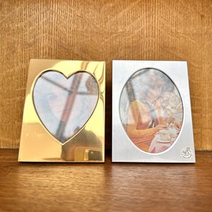 May include: Two metal picture frames. One is gold-coloured and heart-shaped, the other is silver-coloured and oval. Both frames have pictures inside and are displayed on a wooden surface.