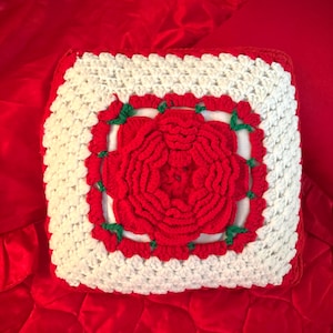 Peut inclure: Un coussin carré fait à la main au crochet, avec un grand motif de rose rouge au centre. La rose est entourée d'une bordure blanche et agrémentée de détails verts. Le coussin est posé sur un fond rouge.
