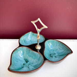 Peut inclure: Un plat de service à trois compartiments avec un vernis brillant turquoise et brun foncé. Le bol en céramique présente une poignée centrale en métal doré avec une boucle en forme de losange au sommet, conçue pour transporter le plateau.