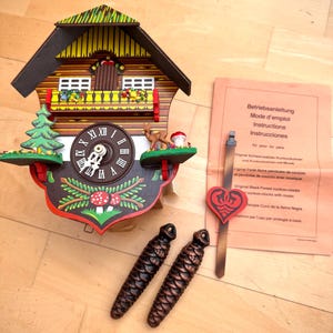 May include: A traditional cuckoo clock with a wooden house design, featuring a clock face, decorative elements, and two pinecone weights. The clock has a brown, red, yellow, and green colour scheme. An instruction sheet is also included.