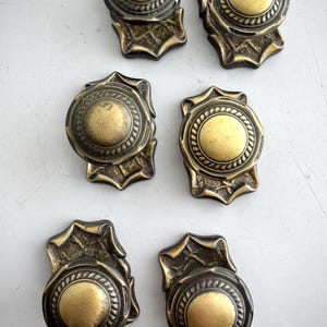 May include: Six antique brass drawer pulls, each with a circular centre and decorative border. The pulls have a shield-like base with ornate detailing. The brass has a warm, aged patina.