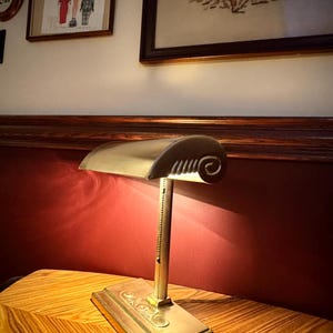 May include: A vintage brass desk lamp with a curved shade and adjustable arm. The lamp features a rectangular base with ornate detailing and a decorative scroll design on the shade. The lamp is a warm gold color and is set against a red background.