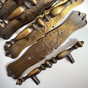May include: Antique bronze cabinet pulls with a textured finish. The pulls have a decorative design with a central, rope-like detail and a curved handle. The hardware is designed for cabinets and drawers.