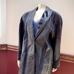May include: A black leather jacket featuring contrasting dark blue suede panels, a wide lapel collar with decorative stitching, long sleeves, and a single button closure at the front. The jacket is displayed on a mannequin over a blue garment.