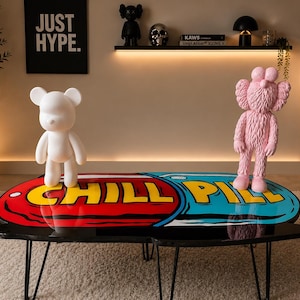 May include: A black coffee table with a colorful pill-shaped design that reads "CHILL PILL" in yellow. A white bear-shaped figurine stands on the left, and a pink figure with crossed-out eyes is on the right.
