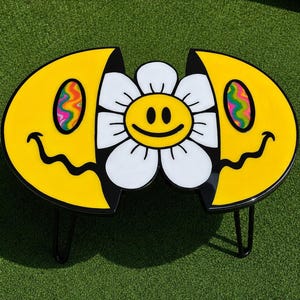 Könnte beinhalten: Ein Couchtisch mit einem geteilten gelben Smiley-Design, wellenförmigen schwarzen Mündern und psychedelischen Wirbelaugen. Eine zentrale weiße Blume mit einem gelben Smiley-Gesicht in der Mitte sitzt zwischen den beiden Hälften.