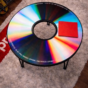 May include: A circular coffee table crafted from a repurposed CD, with a black metal frame. The table's surface displays a vibrant, iridescent pattern. A small red square sits on the table. It rests on a fluffy, white rug.