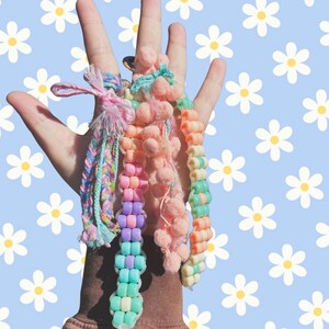 May include: A collection of four handmade bracelets featuring a variety of textures and colors. One bracelet is a braided multi-colored yarn strand, another is made of pastel plastic beads, one features soft pink pom-poms, and one has pastel beads.