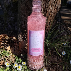 May include: A pink, glitter-covered glass bottle with a label that reads "Aphrodite's Treasures." The bottle is filled with pink beads and sealed with pink wax. The bottle is surrounded by daisies and greenery.