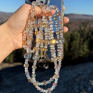 May include: A clear phone case with a mirrored back and a multi-strand beaded phone strap with iridescent beads in shades of clear, gold, and pink.