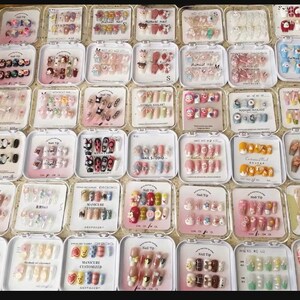 May include: A collection of various press-on nail sets in clear plastic cases. Designs include floral patterns, cartoon characters, and solid colors. The nails are in different shapes and sizes, offering a variety of manicure options.