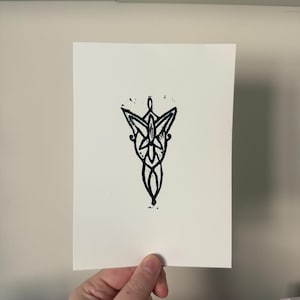 May include: A black and white linocut print of a stylized, symmetrical design. The artwork features a central, elongated shape with intricate details, including a floral or leaf-like element at the top and a series of interconnected lines.