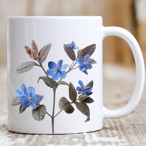 Puede incluir: Taza de cerámica blanca con un diseño de flores azules. Las flores son azules con un centro blanco y tienen hojas verdes.