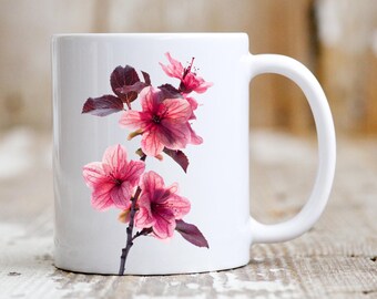 Könnte beinhalten: Weiße Keramiktasse mit einem rosa-weißen Blumendesign. Die Blumen sind zart und haben eine weiche, aquarellartige Optik.