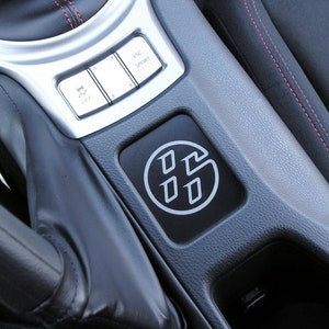 May include: Close-up of a car's center console. Features a black panel with a white logo, silver trim, and buttons labeled "VSC SPORT" and "OFF". Red stitching accents the black interior.