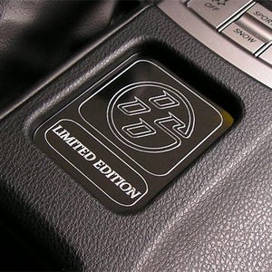 May include: A black rectangular automotive interior emblem featuring a circular logo with stylized letters and the text LIMITED EDITION in white outline. The emblem is set into a textured black plastic console surface near silver control buttons.