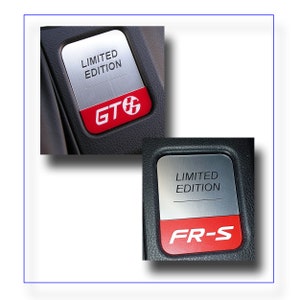 May include: Two rectangular metal car emblems. The top portion is brushed silver with the words "LIMITED EDITION" in black. The bottom portion is red with the letters "GT" and a symbol in white. The second emblem has the letters "FR-S" in white.