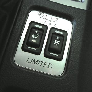 May include: A brushed metal panel with two black buttons featuring seat heater icons. The panel also includes a gear shift pattern and the word "LIMITED". The panel is set in a black textured surface.