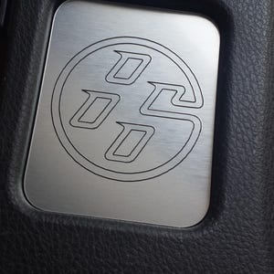 May include: A brushed metal plate with a black outline of a stylized "86" logo, set against a black, textured background. The plate is rectangular with rounded corners, and the logo is centered.