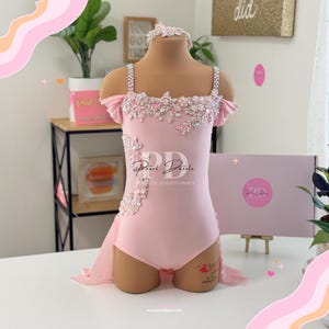 以下が含まれることがあります： 花柄とラインストーンをあしらったピンクのダンスコスチューム。コスチュームはハート型のネックラインとフィットしたボディス。スカートは流れるような重ね着のチュール。コスチュームはマネキンに着せられています。ボディスには「Pearl Dazzle Dance Costumes」の文字が書かれています。