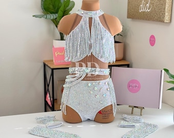 May include: A white dance costume with a fringe top, a bedazzled skirt, and matching arm cuffs. The costume is on a white mannequin. The text "Savich for Dance Jamaica" is visible on the skirt.