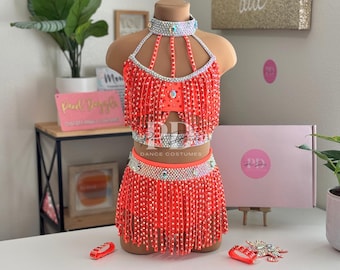 May include: A vibrant neon orange dance costume, comprising a halter top and skirt, both featuring beaded fringe. The top and skirt are embellished with rhinestones, and a matching choker is included. The words "DANCE COSTUMES" are visible.