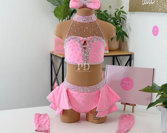May include: A pink dance costume set with a matching headband, choker, and arm cuffs. The set features a bedazzled halter top, a skirt with a ruffled hem, and is embellished with rhinestones. The costume is displayed on a mannequin.