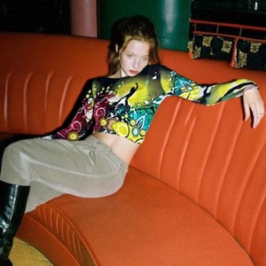 Op de afbeelding: Een vrouw draagt een kleurrijk, abstract geprint lang mouw crop top en een grijze broek en zit op een oranje bank.