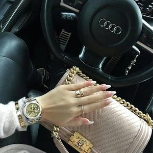 Puede incluir: Un reloj de pulsera dorado y plateado se lleva en la muñeca. Un bolso acolchado beige con una correa de cadena dorada y un cierre está en primer plano. El volante de un coche está en el fondo.