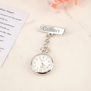 Op de afbeelding: Een zilveren verpleegster zakhorloge met de naam "Gillian" op een rechthoekig label. Het horloge heeft een witte wijzerplaat met zwarte cijfers en wijzers, en een rode secondewijzer. Een ketting verbindt het horloge met het label.