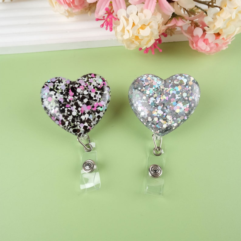 May include: Two heart-shaped badge holders with glitter. One is black with pink and white glitter, the other is silver with iridescent glitter.