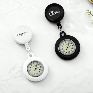 May include: Two retractable badge reel watches, one white and one black. The white watch has the name "Henry" on the reel, and the black watch has "Clare". Both watches have a white clock face with black numbers and hands.