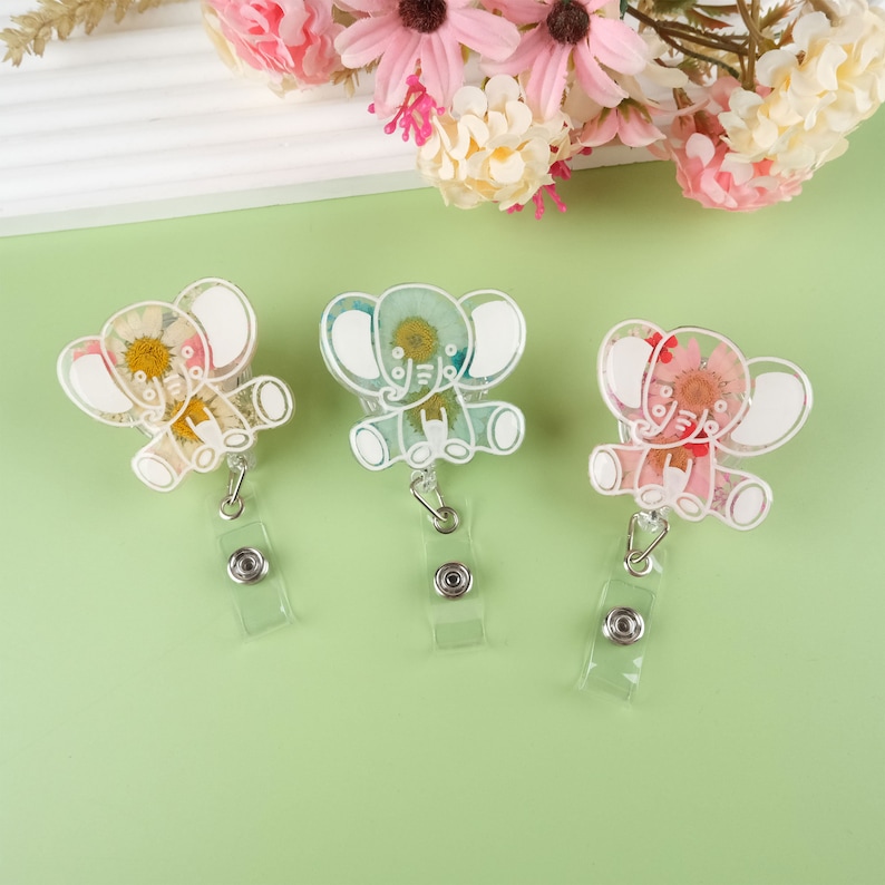 May include: Three clear plastic elephant-shaped badge holders with dried flowers inside. The elephants are white with a light blue, green, and pink flower design. Each holder has a clear plastic retractable badge reel.