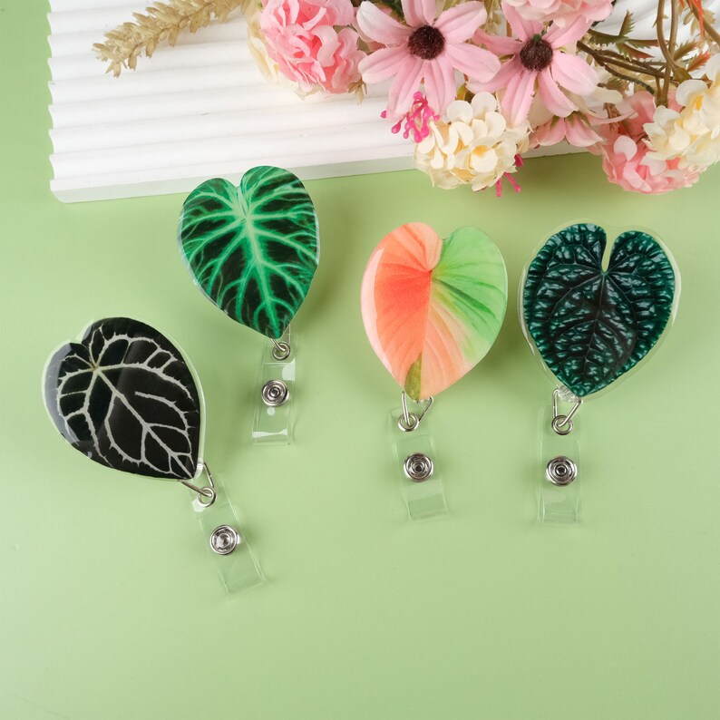 May include: Four different leaf-shaped badge holders with clear retractable cords. The leaves are green, black, pink, and green with a white stripe.