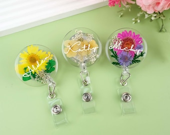 May include: Three clear round resin badge holders with a retractable reel. Each holder features pressed flowers and a name in white script. The names are Alden, Kathy, and Micky.