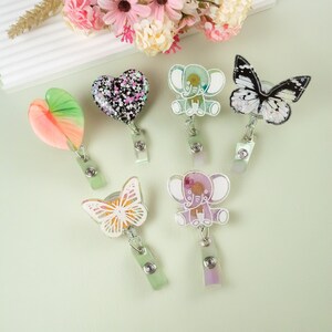 May include: Six colorful resin badge holders with different designs. The designs include a heart, two elephants, and two butterflies. The badge holders are attached to retractable badge reels.