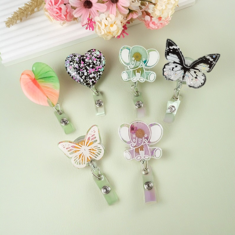 May include: Six colorful resin badge holders with different designs. The designs include a heart, two elephants, and two butterflies. The badge holders are attached to retractable badge reels.