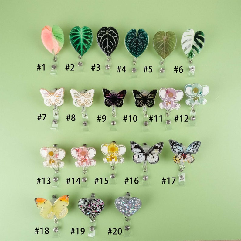 May include: Twenty different badge reel charms featuring various designs, including butterflies, elephants, hearts, and plants. The charms are made of clear acrylic and have a silver metal clip.