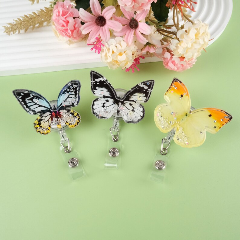 May include: Three butterfly-shaped retractable badge holders with glitter accents. The butterflies are black and white, yellow and orange, and blue and green.