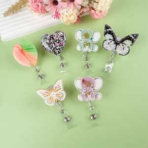May include: Six colorful resin badge reels with different designs. The designs include a heart, two elephants, and two butterflies. The reels are attached to clear plastic retractable cords.