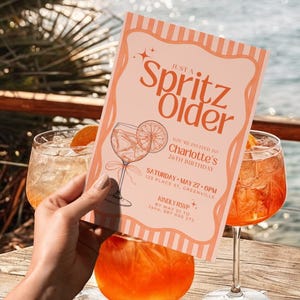以下が含まれることがあります： 「Just a Spritz Older」の文字が入った桃色の招待状が、2つのカクテルの前に掲げられています。招待状には、シャーロットの26歳の誕生日の詳細が記載されており、カクテルグラスの絵が描かれています。背景には木のテーブルと水があります。