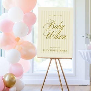 Gender-neutral yellow baby shower welcome sign displayed on a wooden easel at a styled shower setup with balloons and florals — editable A0 Canva template with soft vertical stripes.