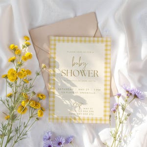 May include: A baby shower invitation with a yellow gingham border and gold lettering. The invitation includes the text "baby shower" and event details. Yellow and purple flowers and a beige envelope are arranged on white fabric.