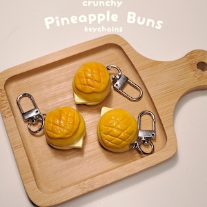 May include: Three yellow pineapple bun keychains with silver clasps rest on a wooden tray. The buns have a textured, grid-like pattern on top and a white filling. The text "crunchy Pineapple Buns keychains" is above.