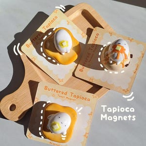Puede incluir: Tres imanes Buttered Tapioca en tarjetas de presentación individuales. Dos imanes muestran un personaje blanco sobre una forma de tostada dorada con un cuadrado de mantequilla amarillo. Un imán muestra un personaje blanco sobre un panqueque.