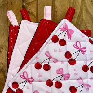 May include: A collection of quilted pot holders with pink hanging loops. The pot holders display patterns such as red with white hearts, solid red, and white with red cherries and pink bows. They are arranged on a wooden surface.